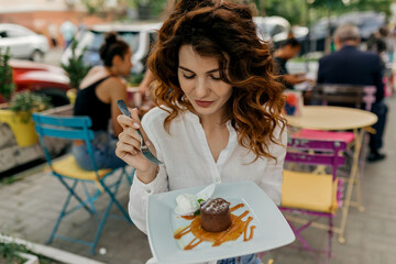 Outdoor portrait of charming pretty woman with curly hair dressed white shirt eating dessert on summer terrace