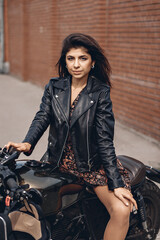 Obraz na płótnie Canvas Portrait of a gorgeous tanned attractive woman wearing a black leather jacket and dress and sitting on a black motorcycle posing outdoors against a brick wall. Speed ​​concept