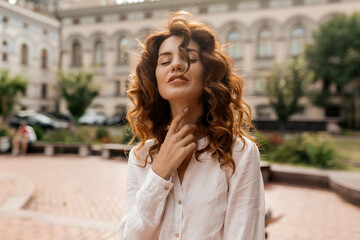 Attractive stylish woman with red curly hair dressed white shirt touching her neck with closed eyes and smiling on background of old city streets