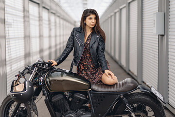 Fototapeta na wymiar Portrait of a bright and daring adult model in a leather jacket and dress posing next to a black motorcycle and looking straight into the distance against the backdrop of white walls. 