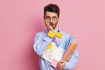 Shocked unshaven man gazes surprisingly keeps hand on cheek holds paper documents with written information data wears formal clothes isolated over pink background has deadline for organizing job