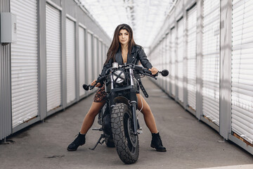 Naklejka na ściany i meble Lovely young and brutal biker movel in a leather jacket posing on a black retro motorcycle holding the steering wheel with her hands and looking into the distance. Extreme concept