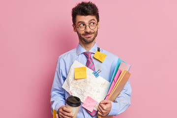 Positive thoughtful office worker has happy look aside thinks how to prepare financial project wears formal shirt with stickers drinks coffee from disposable cup isolated over pink background