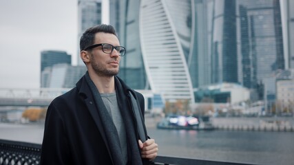 Man in black coat and eyeglasses walking on background of business skyscrapers in Moscow, slow...