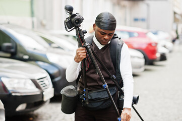 Fototapeta na wymiar Young professional african american videographer holding professional camera with tripod pro equipment. Afro cameraman wearing black duraq making a videos.