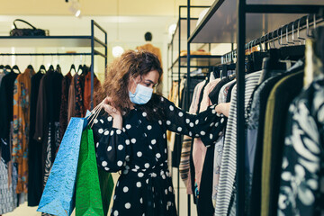 Young beautiful woman in a protective mask on her face, chooses clothes in the store, makes safe...