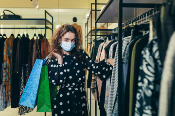 Young beautiful woman in a protective mask on her face, chooses clothes in the store, makes safe...