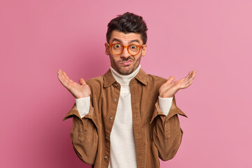 Hesitant clueless unshaven man spreads palms and faces difficult choice looks confused wears transparent glasses and brown shirt poses against rosy background. Uncertain confused European guy - obrazy, fototapety, plakaty