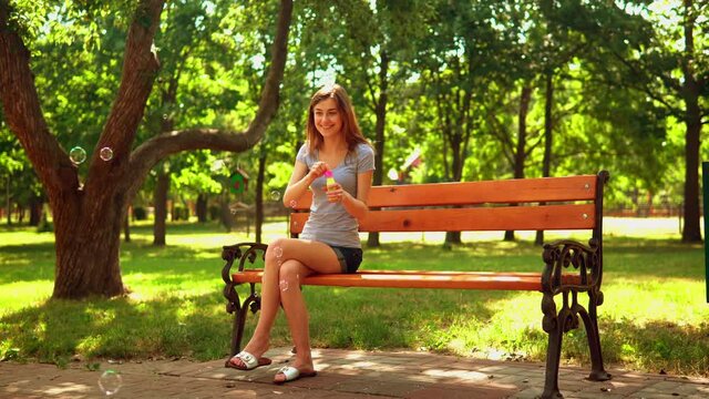 slim young woman in denim shorts sitting on the bench have fun laughing supplying soap bubbles on the background spring garden