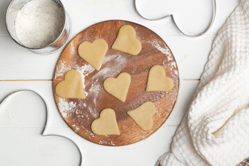 A heart shaped cutout mold and dough cut cookies next to a towel and wooden spoons and a mug for sifting flour.