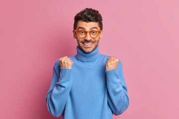 Positive unshaven man clenches fists celebrates something with joy rejoices good results of his work smiles broadly wears casual blue turtleneck isolated over pink wall. Yeah finally achieved goal