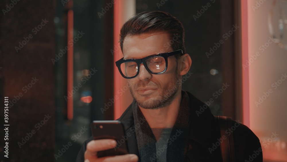 Fototapete Man in eyeglasses with phone in hand on background of building with lights on. Handheld night shot of caucasian man texting in phone near business building with cars