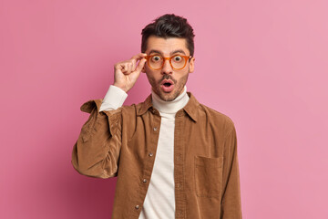 Amazed emotional unshaven guy stares through glasses shocked to hear amazing news keeps mouth...