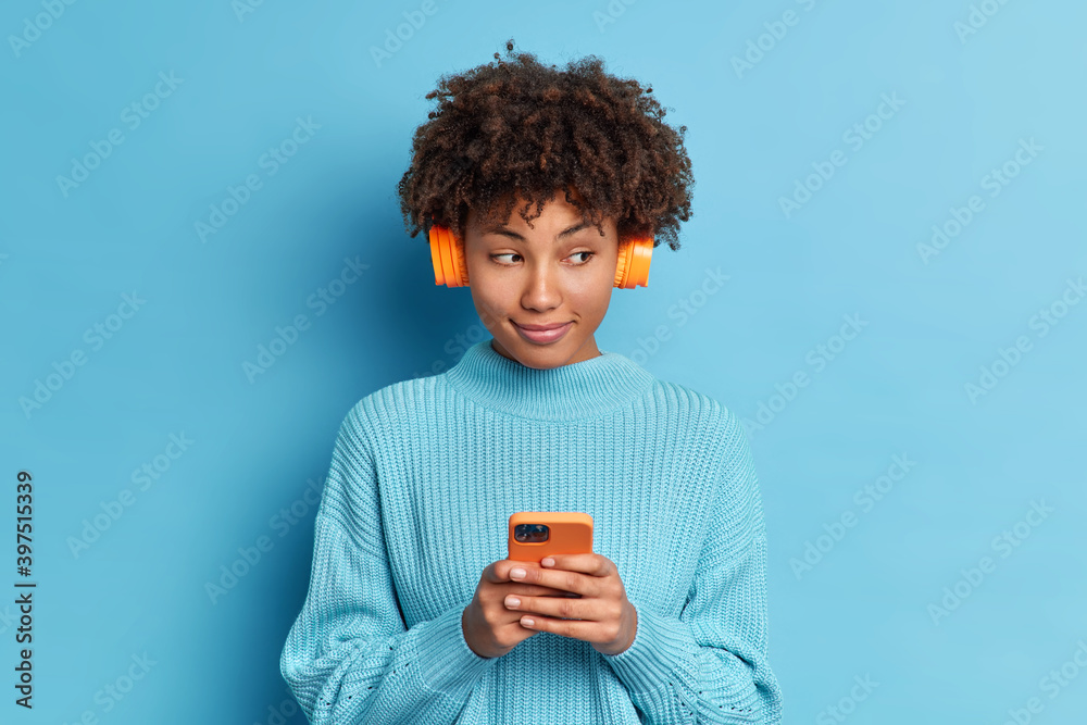 Wall mural Photo of beautiful dark skinned girl with pensive expression listens favorite track in headphones holds modern cellular and sends text messages wears casual jumper isolated over blue studio wall - Wall murals