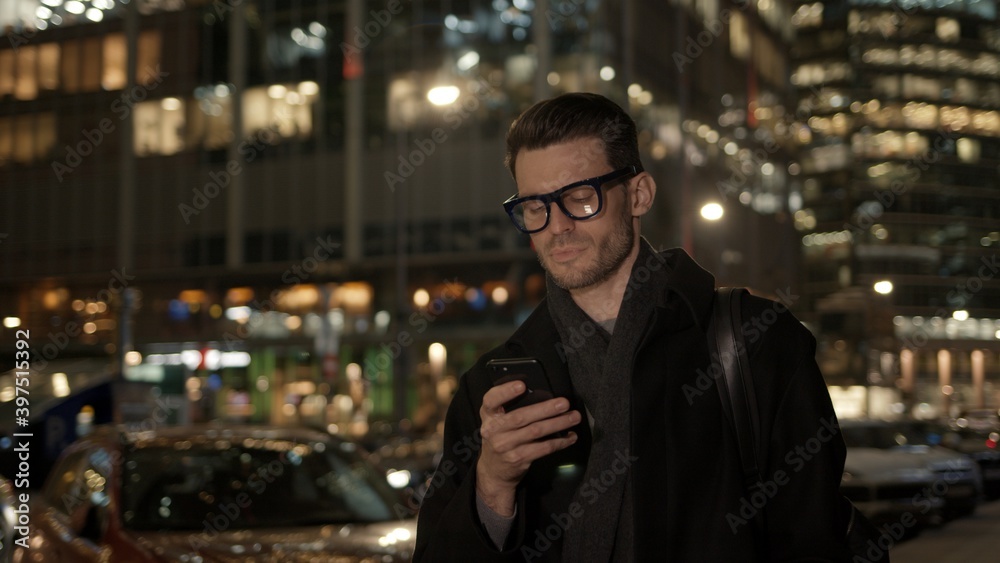 Fototapete Man in eyeglasses with phone in hand on background of building with lights on. Gimbal night shot of caucasian man texting in phone near business building