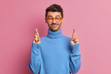 Young cheerful adult man with bristle crosses fingers believes in good luck before important job...