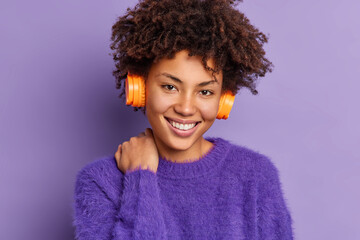 Headshot of good looking curly haired woman with Afro hair smiles gently touches neck looks positively at camera listens audio track via wireless stereo headphones isolated over purple background