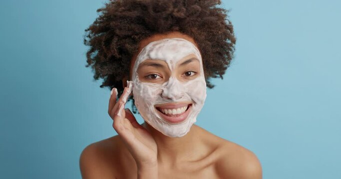Beautiful cheerful Afro American woman applies facial mask touches face gently waits for nice effect after cosmetic product stands indoor isolated on blue background takes care of her complexion