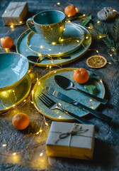 Festive Christmas table setting. Composition from a blue plate, golden cutlery, garland, oranges and gift boxes on brown background, top view, vertical image.