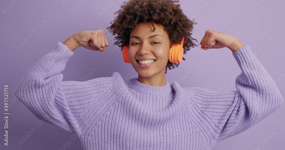 Wall mural Self assured proud curly ethnic woman brags about her strength raises arm and shows muscles wears stereo headphones casual long sleeved jumper isolated over purple background. Look at my biceps