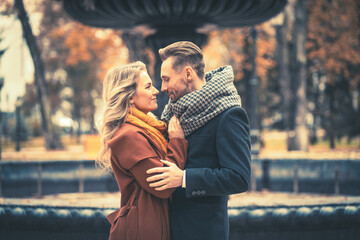 Close up. Loving couple of young people standing embracing and looking at each other near a old fountain in the autumn park wearing autumn coats. Toned photo.  - Powered by Adobe
