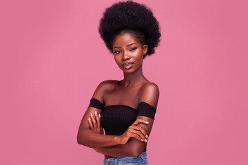Confident African American girl with afro hair standing light smiling with arms folded in black bare shoulder top and blue denim jeans isolated on pink background.  - obrazy, fototapety, plakaty