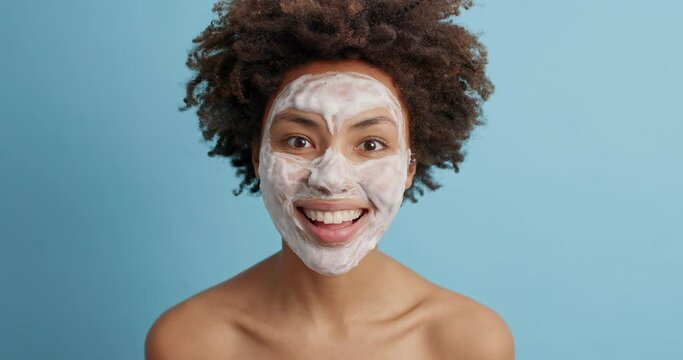 Curly young woman makes funny grimace pouts cheeks has fun wears beauty face mask for healthy smooth skin undergoes cosmetic facial treatment stands bare shoulders isolated on blue background