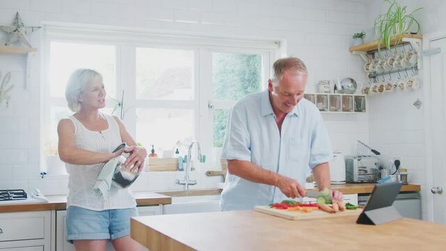 Retired senior couple making meal in kitchen with man following recipe on digital tablet - shot in slow motion