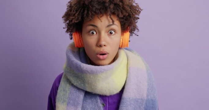 Wow what indredible news. Impressed amazed dark skinned woman looks with great wonder at camera drops jaw from surprisement hears something unbelievable wears stereo headphones scarf around neck