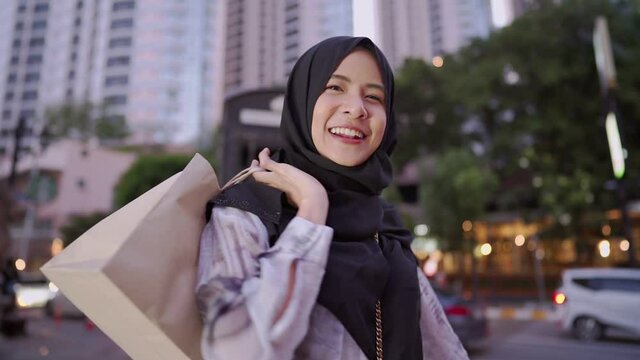 young Asian muslim feel happy showing shopping bags to camera, standing on the street, capitalist consumer spending money, discounts sales products, enjoy shopping at the mall, downtown city lifestyle