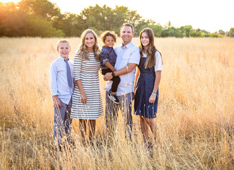 An elegant and beautiful young family portrait outdoors in a grassy field. Smiling and happy diverse group with a mixed race adopted baby boy and two other children