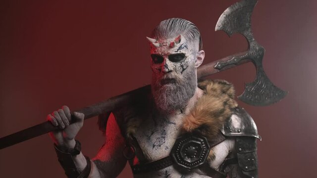 Winter dead warrior with pale skin and beard poses in dark red background with smoke holding his double axe on his shoulder rotating his head and looking around.