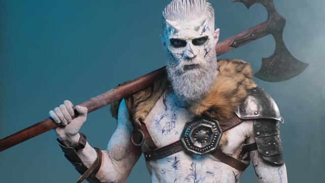 Mysterious and dangerous portrait of a undead northern fighter with pale skin and beard posing in dark blue background with smoke holding his huge axe on his shoulder and looking at camera.