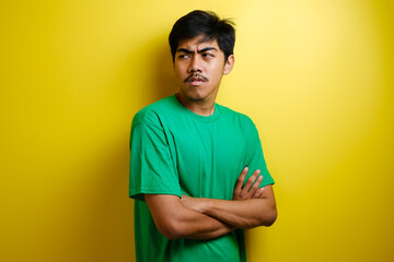 Asian young man wearing yellow t-shirt crossed his arm on chest while looking to his side in anger