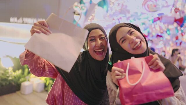Two young asian excited Muslim women holding showing paper shopping bags while standing at shopping center inside modern downtown during the night, new lucky purchase bought on sale with discount 