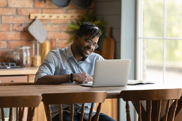 Happy young mixed race man sitting at table, enjoying learning distantly on online courses, using computer at home. Smiling 30s businessman in eyewear improving knowledge, watching webinar lecture.