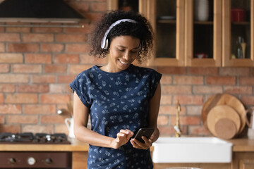 Smiling attractive young african american woman in headphones using mobile music library application, choosing favorite audio, starting preparing food on weekend in kitchen, modern tech concept.
