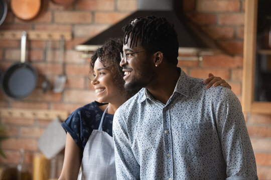 Happy young bonding african american couple looking in distance, daydreaming together in kitchen. Loving millennial mixed race wife and husband visualizing future, looking at same side indoors.
