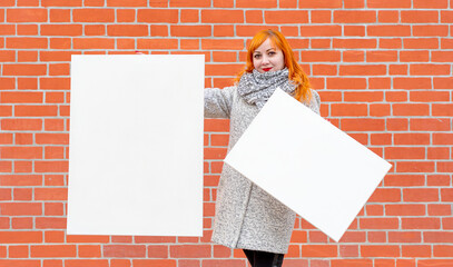 A young red-haired girl stands against the background of a red wall and holds two blank white canvases for painting in front of her