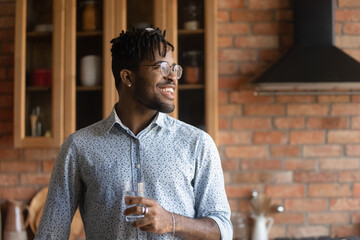 Dreamy young mixed race african man in eyeglasses looking in distance, holding glass of fresh clear water in hands, copy space for advertising text, good daily morning healthcare habit concept. - Powered by Adobe