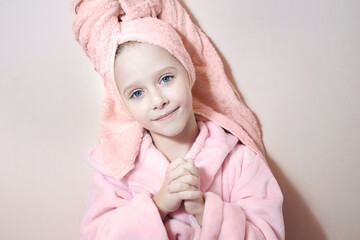 a little girl with blue eyes in a pink robe and a pink towel on her head after a bath smiles and looks into the frame. on a peach background