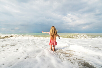 a little girl in a pink dress stands facing the sea and looks at the foaming waves