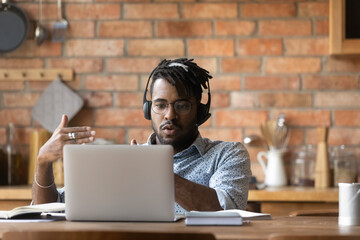 Serious young african ethnicity man in eyeglasses and modern wireless headset looking at computer...
