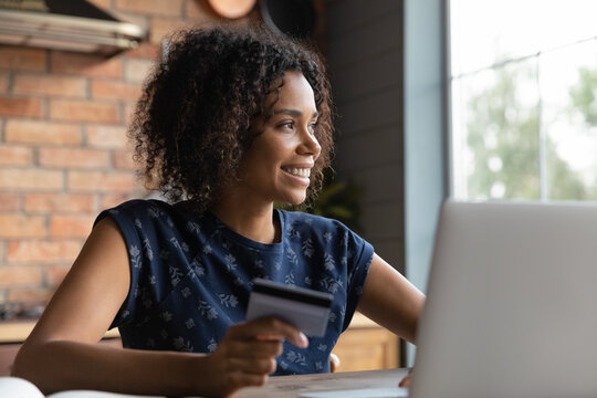 Smiling Young African American Woman Holding Plastic Bank Card In Hands, Planning Products Purchase In Internet Store, Looking In Distance, Dreaming Of Sale Season, Planning Buying Gifts Online.