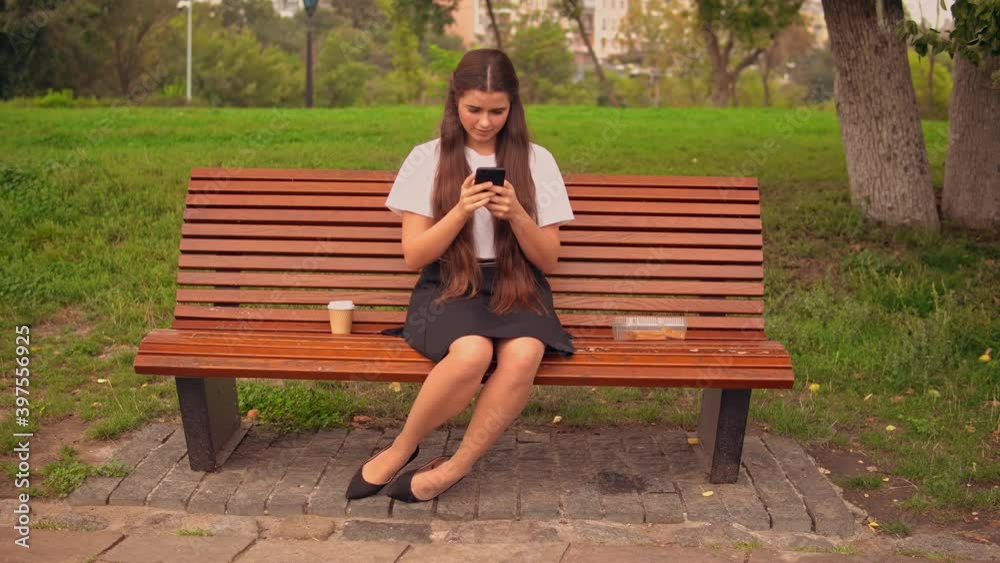 Poster break for lunch student sitting on the bench typing message or scrolling news feed on smartphone drinking coffee to go summer season outdoors