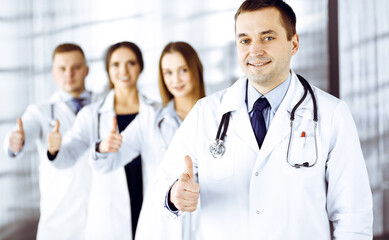 Group of professional doctors are standing as a team with thumbs up in a hospital office, ready to help their patients. Medical help, insurance in health care and medicine concept