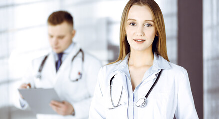 Fototapeta premium Professional beautiful woman-doctor with a stethoscope is standing with crossed arms in a clinic. Young doctors at work in a hospital. Medicine and healthcare concept