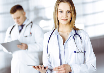 Fototapeta premium Professional woman doctor with a stethoscope is using a computer tablet, while she is standing together with her colleague in a clinic. Young doctors at work in a hospital. Medicine and healthcare