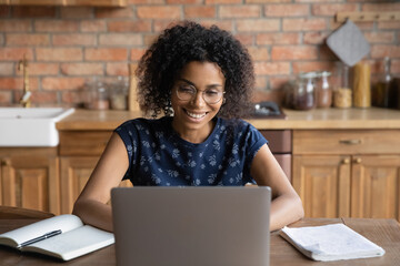 Happy young african american woman in eyeglasses working on computer in kitchen, smiling millennial mixed race female freelancer enjoying using laptop application, web surfing online at home office.