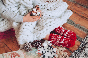 Little girl sitting on the floor in red festive Christmas socks under a chunky knit blanket holding...